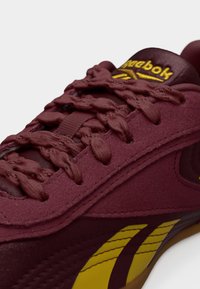 Μαρόν αθλητικό παπούτσι Reebok με κίτρινο λογότυπο, λεπτομερείς κορδόνια και υφασμάτινη υφή, κοντινή λήψη σε λευκό φόντο.