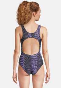 Vrouw met krullend haar in een paars en zwart gestreept badpak met een grote open rug, staand met de rug naar de camera tegen een witte achtergrond.