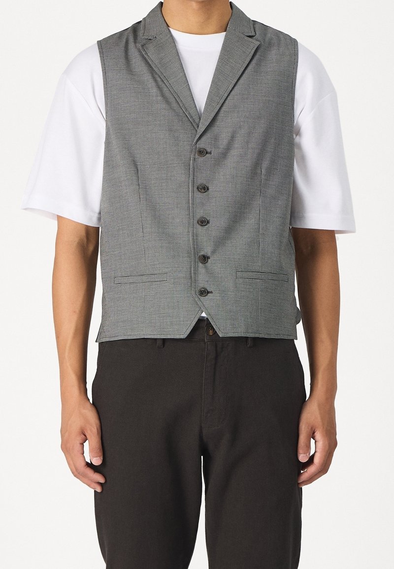 Gilet grigio a quadri con un design su misura, cinque bottoni e rever a punta. Indossato sopra una t-shirt bianca, abbinato a pantaloni scuri.