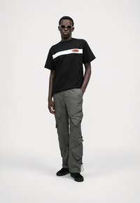 T-shirt nera con striscia bianca e logo, abbinata a pantaloni cargo verde oliva con molteplici tasche e dettagli con cordoncino. Scarpe nere.