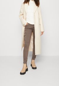 Beżowy długi płaszcz nałożony na biały golf, w połączeniu z brązowymi spodniami cargo i beżowymi botkami za kostkę z masywnymi czarnymi podeszwami.