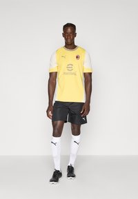 Maglia da football corta giallo e bianca con un logo, abbinata a pantaloni neri e calzettoni bianchi. Il giocatore indossa scarpini neri.