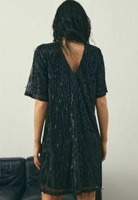 Schwarzes Kleid mit vertikalen Pailletten, V-Ausschnitt, kurzen Ärmeln und Fransenbordüre. Glatte Textur und lockerer Sitz.