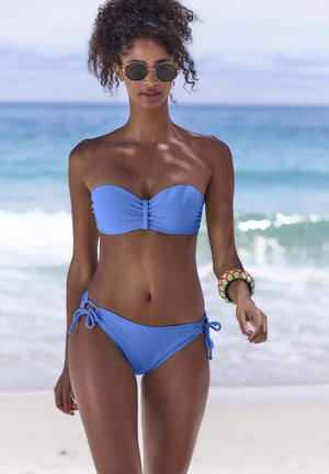 Mujer con bikini azul bandeau y pulsera colorida camina por la playa de arena con olas del océano al fondo.