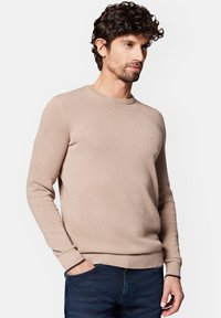 Beżowy sweter w robocie z okrągłym dekoltem, teksturowanym materiałem oraz prążkowanymi mankietami i dołem. Mała emblemat na piersi, zestawiony z ciemnymi dżinsami.