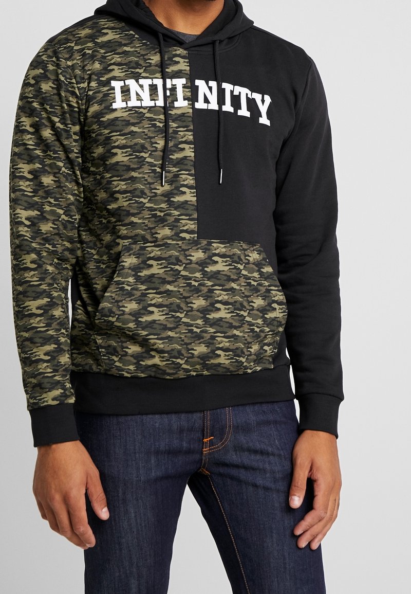 Homme portant un sweat à capuche noir et camouflage avec le texte blanc "INFINITY", associé à un jean bleu foncé.