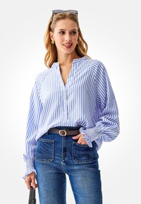 Blauw-wit gestreepte blouse met kraag, knoopsluiting aan de voorkant en elastische manchetten. Gecombineerd met donkerblauwe jeans met hoge taille en een zwarte riem.