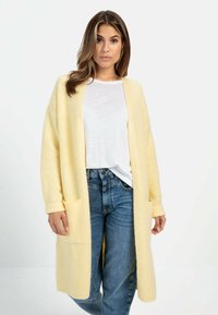 Geel gebreide cardigan met lange mouwen en zakken, gecombineerd met een wit T-shirt en blauwe jeans. Textuurrijke oppervlakte en een ontspannen pasvorm.