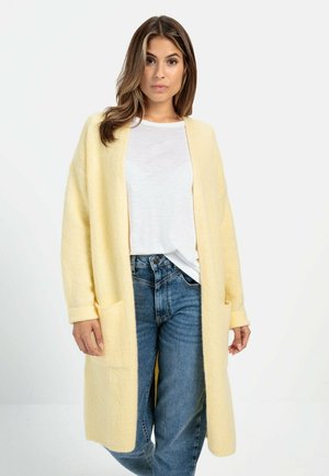 Gul strikket cardigan med lange ærmer og lommer, parret med en hvid T-shirt og blå jeans. Struktureret overflade og afslappet pasform.