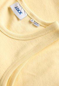 T-shirt jaune clair avec un tissu texturé, un col rond et des détails côtelés. Étiquette de la marque visible indiquant la taille "S" et le pays de fabrication.