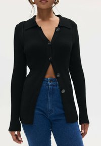 Czarny żeberkowany kardigan z kołnierzykiem, zapięciem na guziki i długimi rękawami. Noszony na wysoko-wzorzystych niebieskich dżinsach, odsłaniający strefę brzucha.