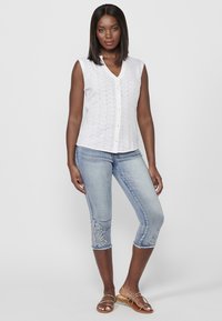 Chemisier sans manches blanc à boutons avec des détails en broderie perforée, associé à un jean capri bleu clair avec de la broderie florale sur la jambe gauche. Sandales beiges.