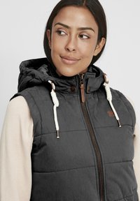 Svart vadderad väst med huva, med en brun dragkedja, snörning med stopp och en liten brun logotyp på bröstet.