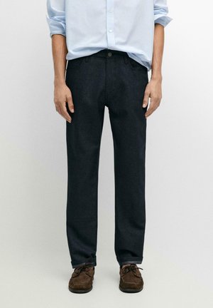 Mężczyzna w ciemnych prostych dżinsach w połączeniu z brązowymi sznurowanymi butami i jasnoniebieską koszulą w paski z podwiniętymi rękawami.