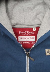 Hættetrøje med en marineblå yderside og grå foring, udstyret med lynlås, en rektangulær lappesøm og et rødt stofmærke indeni.