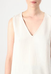 Sleeveless top in a light beige fabric with a subtle texture, featuring a deep V-neckline and clean, minimal design.