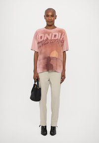 T-shirt oversize rose avec un graphique de coucher de soleil et le texte "DONDUI made in Italy." Assorti avec un pantalon beige et un sac noir, bottes à talons noirs.