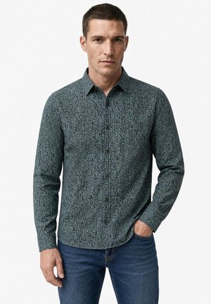 Hombre de pie con una mano en el bolsillo, vistiendo una camisa de manga larga con botones con un pequeño patrón geométrico y vaqueros azules.