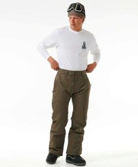 Un snowboardeur portant une chemise blanche à manches longues, un pantalon vert olive, des boots noires et un bonnet marron avec des lunettes de protection reposant dessus.