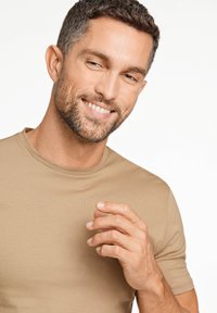 Beżowa dopasowana koszulka z okrągłym dekoltem, wykonana z gładkiego materiału. Model ma krótkie ciemne włosy i się uśmiecha, trzymając rękę blisko klatki piersiowej.