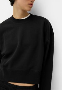 Črna skrajšana trenirka iz mehkega materiala, z okroglim izrezom, rebrastimi manšetami in sproščenim krojem, z vidnimi šivi.