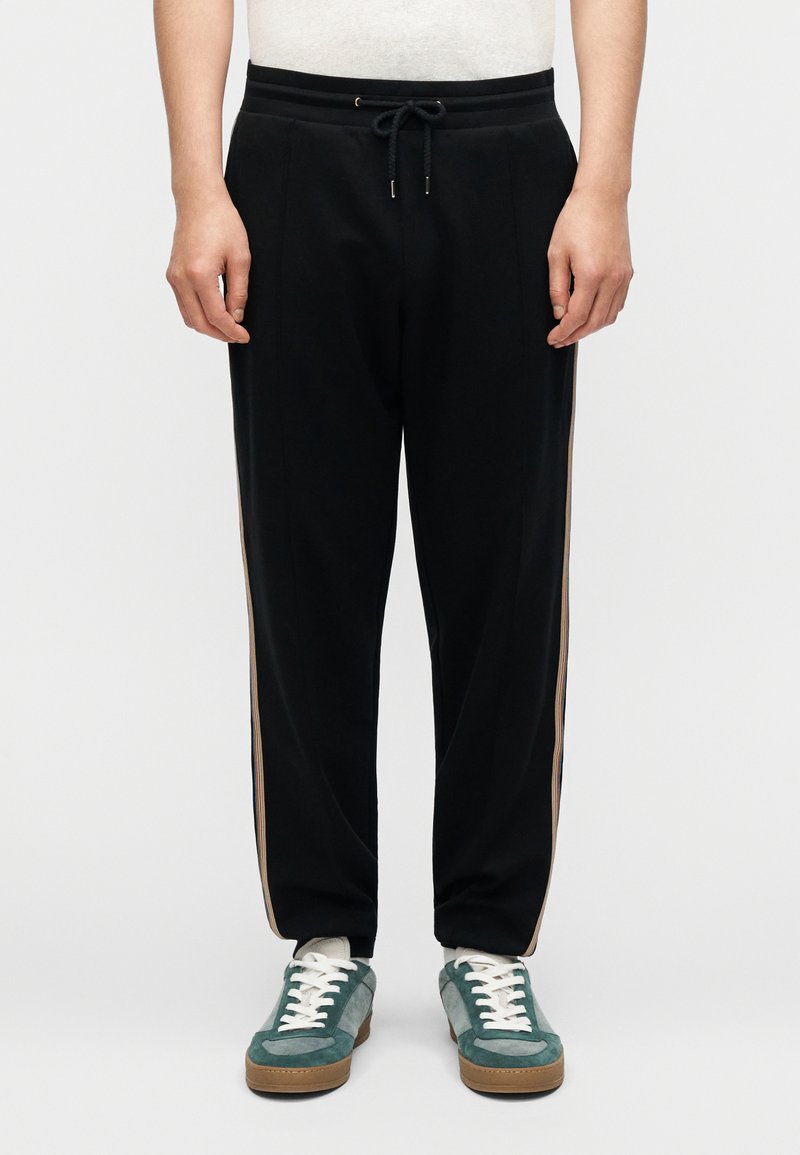 Μαύρο αθλητικό παντελόνι με κορδόνι στη μέση, διακοσμημένο με πλαϊνές ρίγες σε μπεζ χρώμα. Το ύφασμα φαίνεται λείο, με άνετη εφαρμογή. Φορεμένο με πράσινα αθλητικά παπούτσια.