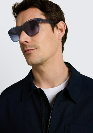 Man draagt donkerblauwe doorschijnende vierkante zonnebril, een marineblauwe jas met zichtbare rits en een wit overhemd tegen een grijze achtergrond.