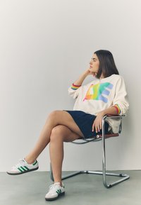 Jeune femme aux cheveux foncés assise sur une chaise en métal, portant un pull blanc avec "LOVE" aux couleurs de l’arc-en-ciel et des baskets blanches avec des rayures vertes.