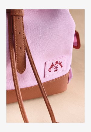 Zaino in tessuto rosa con cinghie e base in pelle marrone, con una toppa cucita con testo rosso in corsivo e un piccolo disegno a nastro.