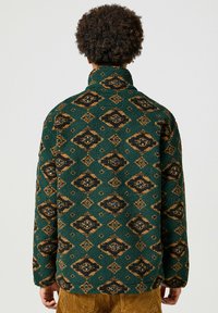 Chaqueta de forro polar verde con cuello alto, que presenta un patrón de diamantes en negro y marrón. Textura suave con un corte holgado y mangas largas.