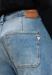 Ανοιχτό μπλε τζιν παντελόνι με πίσω τσέπη που έχει μια μικρή ροζ καρδιά ραμμένη λεπτομέρεια και καφέ ετικέτα της μάρκας MOS MOSH στη μέση.