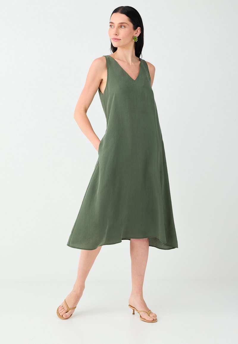 Femme portant une robe midi olive sans manches avec col en V et poches, associée à des sandales à talons couleur beige, debout devant un fond clair uni.