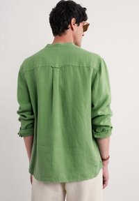 Camicia di lino verde con maniche lunghe, singola piega posteriore e polsini arrotolati. Tessuto morbido, vestibilità casual e orlo leggermente curvato nella parte inferiore.