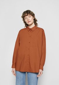 Camisa de botões na cor ferrugem, feita de um tecido suave, com um bolso frontal, colarinho pontiagudo e mangas longas. Combinada com jeans de cor azul clara.