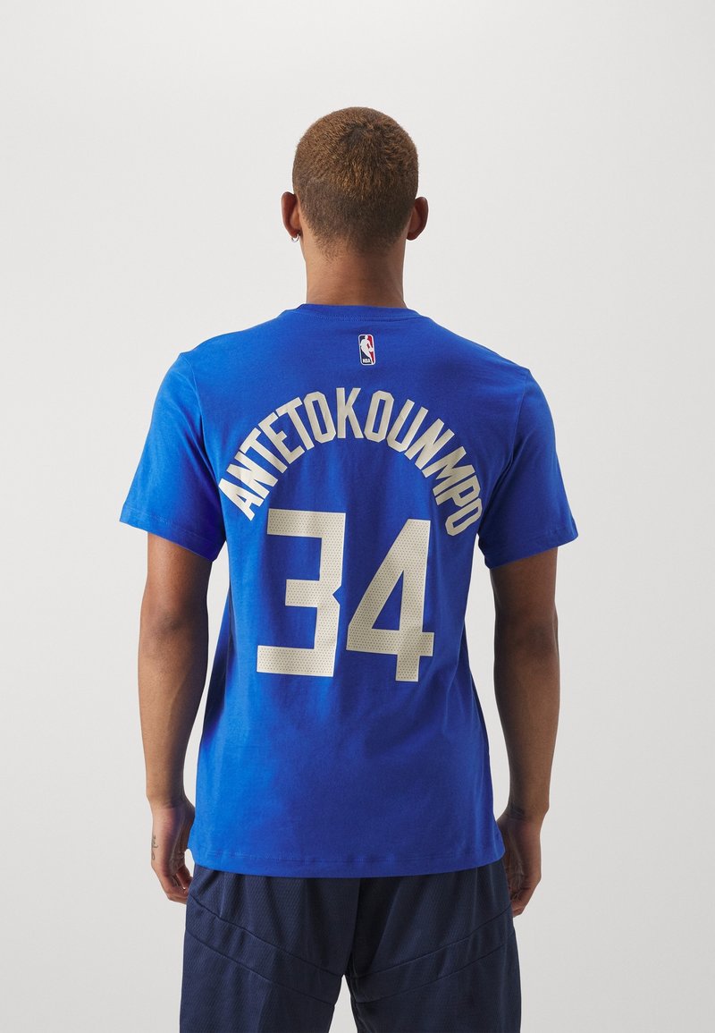 Homme aux cheveux courts portant un maillot bleu de la NBA avec "ANTETOKOUNMPO" et le numéro 34 dans le dos, se tenant devant un fond uni.