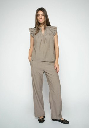 Žena stojaca v svetlohnedom tielku bez rukávov s volánovými ramenami a zodpovedajúcich širokých nohaviciach, ruky čiastočne vreckách, obutá v čiernych balerínach.