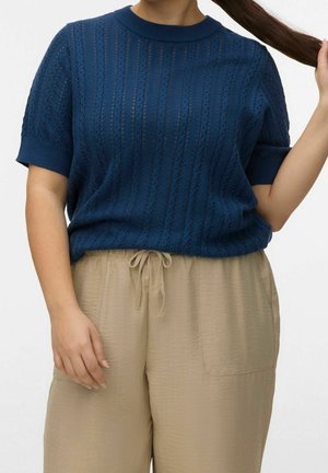 Personne portant un haut tricoté bleu marine à manches courtes et un pantalon beige avec cordon, tenant ses longs cheveux foncés d'une main.