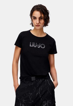 Donna con capelli corti e ricci indossa una t-shirt nera Liu Jo e pantaloni neri con linee astratte bianche, mani in tasca, in piedi contro uno sfondo bianco.