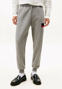 Pantalones de chándal grises con cordón en la cintura, piernas ajustadas, bolsillos laterales y un pequeño parche de logo en rojo y negro en el lateral. Material de mezcla de algodón.