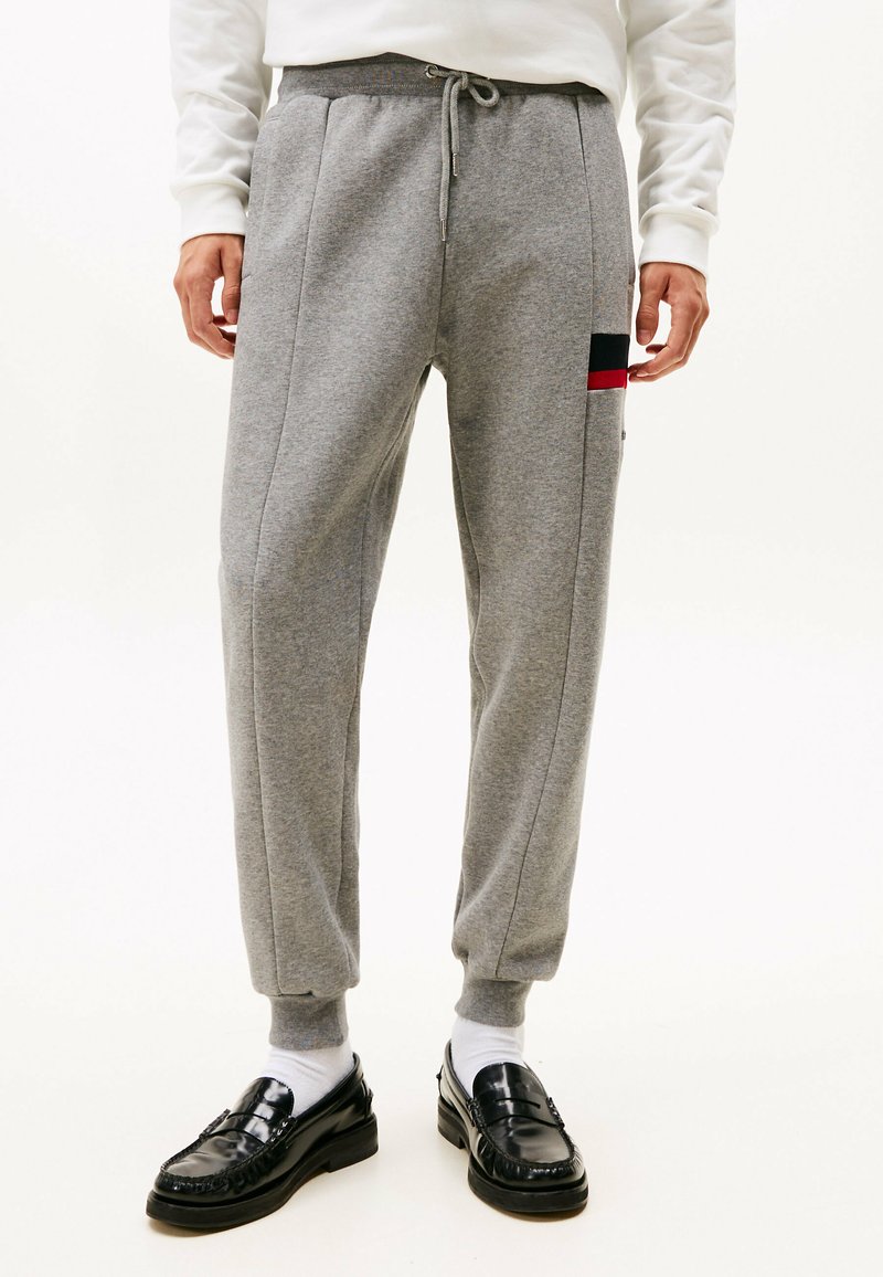 Pantalones de chándal grises con cordón en la cintura, piernas ajustadas, bolsillos laterales y un pequeño parche de logo en rojo y negro en el lateral. Material de mezcla de algodón.