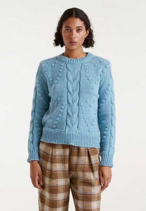 Suéter de punto azul claro con un patrón de trenzas, cuello redondo, y puños y dobladillo acanalados. Llevado sobre unos pantalones de cuadros marrones.