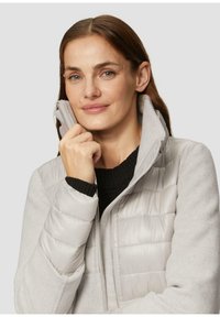 Hellgraue gepolsterte Jacke mit hohem Kragen, aus einem weichen Material gefertigt. Verfügt über einen Reißverschluss auf der Vorderseite und einen kontrastierenden schwarzen Pullover darunter.