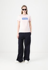 Lys rosa bomull t-skjorte med en blå rektangulær "HUGO" logo, kombinert med vide marineblå bukser og hvite sneakers. Sorte solbriller brukt.