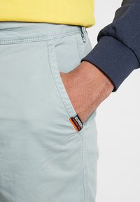 Pantalones de algodón de color azul claro con un bolsillo lateral, que presentan una etiqueta de marca en naranja y negro. El material es suave con un acabado ligeramente texturizado.