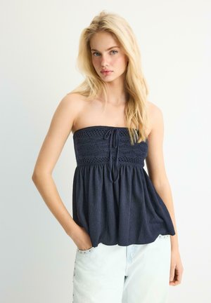 Blondynka z rozpuszczonymi włosami, ubrana w górę bez ramiączek w kolorze granatowym z fakturą oraz jasnoniebieskie dżinsy, z jedną ręką w kieszeni na jasnym tle.