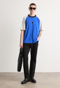 Blue and gray raglan t-shirt with black accents, black pants, and black boots. The model carries a black bag and wears dark sunglasses.