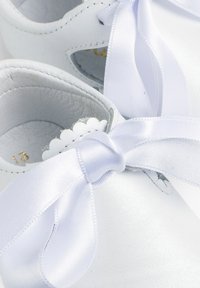 Zapatos de bebé de cuero blanco con acabado brillante, bordes decorativos con ondas y suaves cintas de satén blancas atadas en moños.