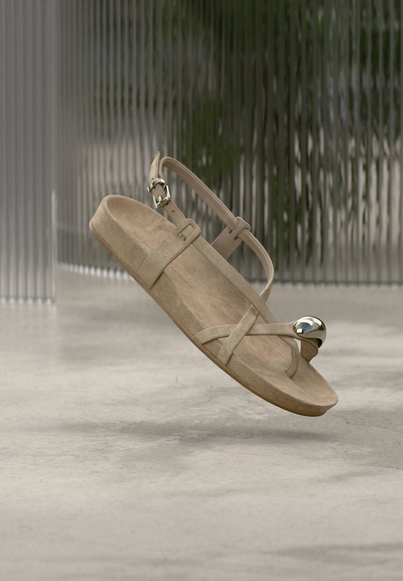 Μπεζ σανδάλι από καστόρι με επίπεδη σόλα, διαθέτει σταυρωτούς ιμάντες και μεταλλική λεπτομέρεια στον αστράγαλο. Μινιμαλιστικός και ελαφρύς σχεδιασμός.
