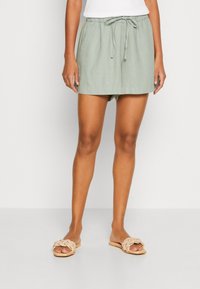 Shorts en lin vert clair avec une taille élastique et un cordon de serrage. Associés à des sandales plates tressées couleur tan.