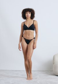Ensemble de bikini noir comprenant un bralette triangulaire avec de fines bretelles et un string taille basse avec des accents en dentelle. Tissu lisse, design minimaliste.